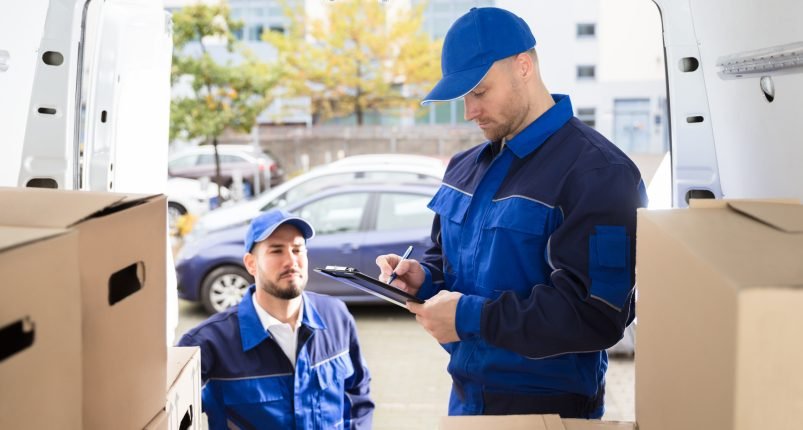 Young Male Mover Looking At His Partner Writing On Clipboard Near Cardboard Boxes